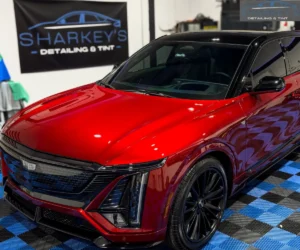 Front-angle view showing the glossy metallic red hood, sleek illuminated grille, and dark multi-spoke wheel of a Cadillac Lyriq